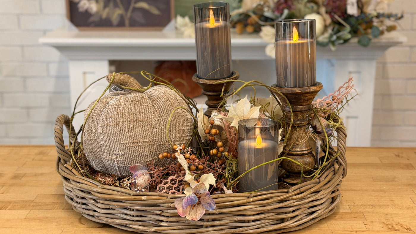 An autumn-themed centerpiece arrangement in a round woven basket, featuring a burlap pumpkin, two tall gray glass candle holders with lit candles, a smaller gray candle, dried flowers, and greenery, displayed on a wooden table in front of a white brick fireplace.