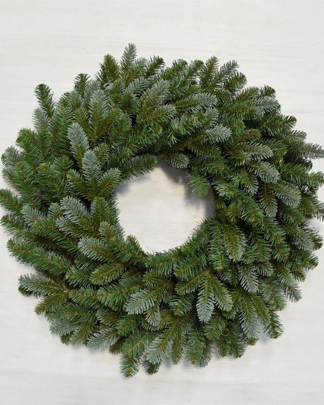 A circular green pine wreath with a frosted, dusty blue-gray finish, photographed from directly above on a light off-white background.