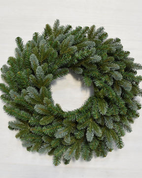 A circular green pine wreath with a frosted, dusty blue-gray finish, photographed from directly above on a light off-white background.