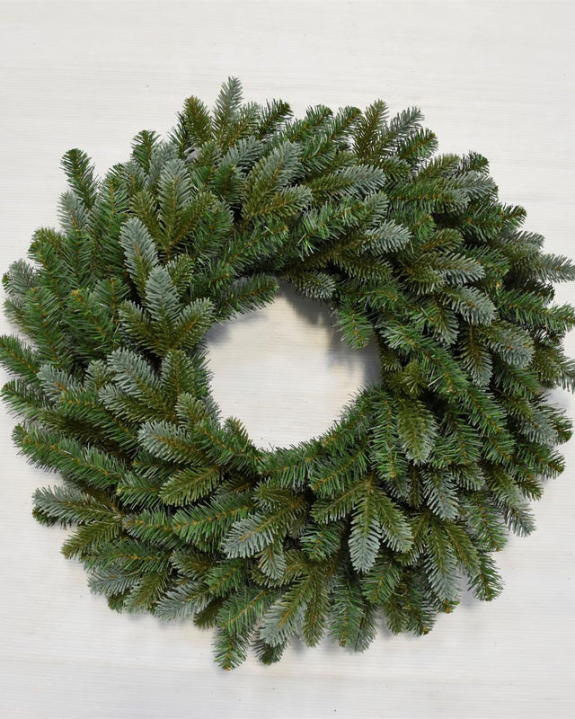 A circular green pine wreath with a frosted, dusty blue-gray finish, photographed from directly above on a light off-white background.