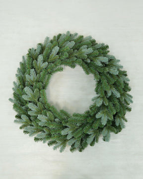 A green artificial pine wreath with a frosted, dusty blue-gray finish, photographed from directly above on a light off-white wooden background.