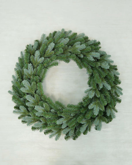 A green artificial pine wreath with a frosted, dusty blue-gray finish, photographed from directly above on a light off-white wooden background.