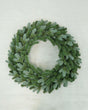 A green artificial pine wreath with a frosted, dusty blue-gray finish, photographed from directly above on a light off-white wooden background.