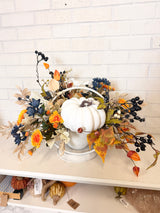 Autumn fall floral arrangement centerpiece featuring a white decorative pumpkin in a white metal basket, surrounded by orange, yellow, blue, and beige faux leaves and berries, displayed on a shelf against a white brick wall.