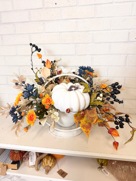 Autumn fall floral arrangement centerpiece featuring a white decorative pumpkin in a white metal basket, surrounded by orange, yellow, blue, and beige faux leaves and berries, displayed on a shelf against a white brick wall.