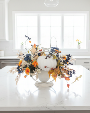 An autumn-themed floral centerpiece with a white pumpkin, blue berries, and orange flowers arranged in a white urn on a white kitchen island, with a bright modern kitchen in the background.