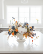 An autumn-themed floral centerpiece with a white pumpkin, blue berries, and orange flowers arranged in a white urn on a white kitchen island, with a bright modern kitchen in the background.
