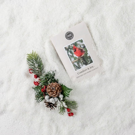 A wax sachet with a Christmas-themed decoration on it, placed on a snowy surface. The sachet is labelled 'Comfort & Joy' with a picture of a red bird on it, indicating it is part of the Cedar Hill Collection.