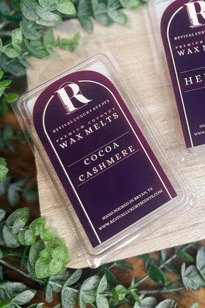 Three wax melts in clear clamshell packaging with dark burgundy labels are arranged on a light wood board surrounded by green eucalyptus leaves. The labels show the brand R and scent names Cocoa Cashmere, Heiress, and Volcanic Ember.